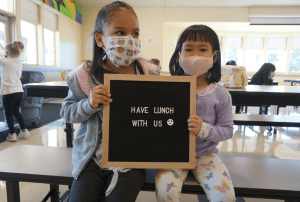 Two students in a cafeteria holding up a "We're Hiring" sign