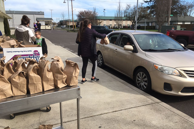 a woman handing a paper bag full of food to a car parked at the curbside