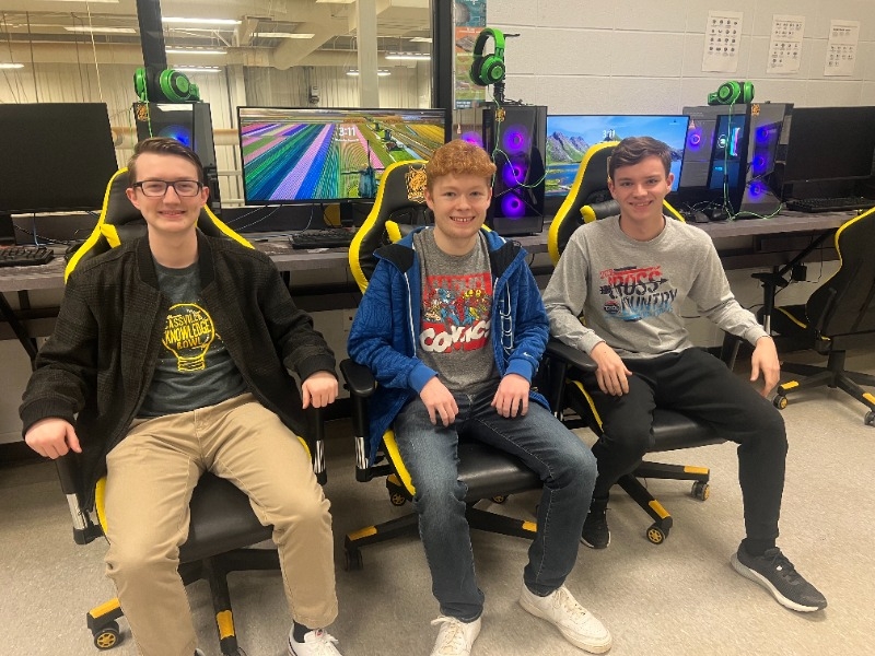 three young men sitting in chairs in front of three computer screens.