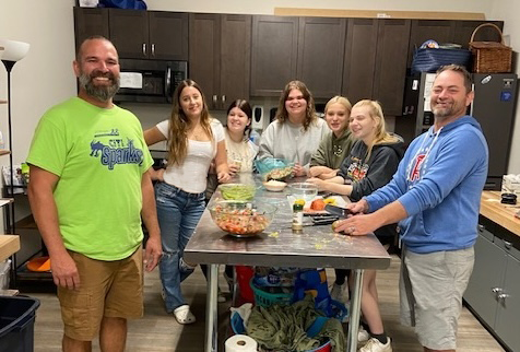 A group of seven smiles while preparing food together in a kitchen, enjoying a collaborative activity.