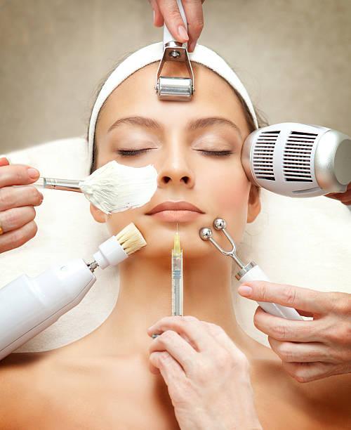 A woman lying down receiving multiple facial cosmetic treatments simultaneously from several hands.