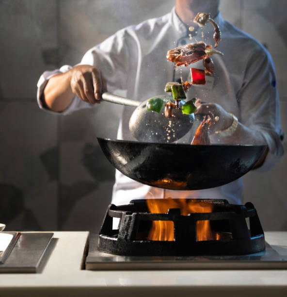 A chef in a white uniform is actively stir-frying food in a black wok over a gas burner with visible flames. 