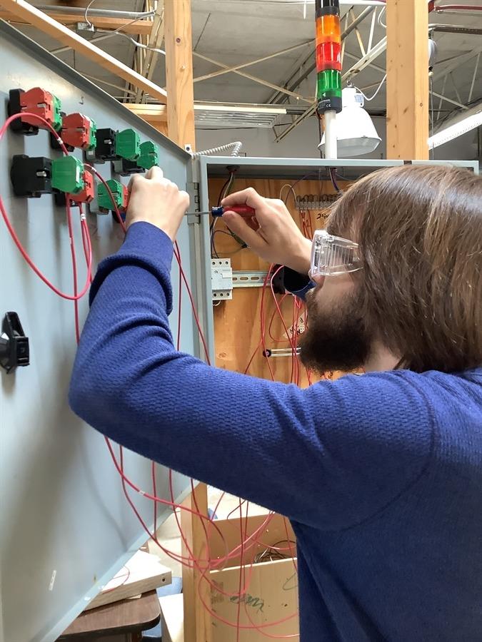 A person wearing safety glasses and a blue long-sleeved shirt is working on electrical wiring inside an open gray electrical panel. 