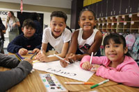 Construyendo Bases Sólidas para el Aprendizaje: Cuatro estudiantes colorean un cartel en un escritorio mientras sonríen a la cámara.