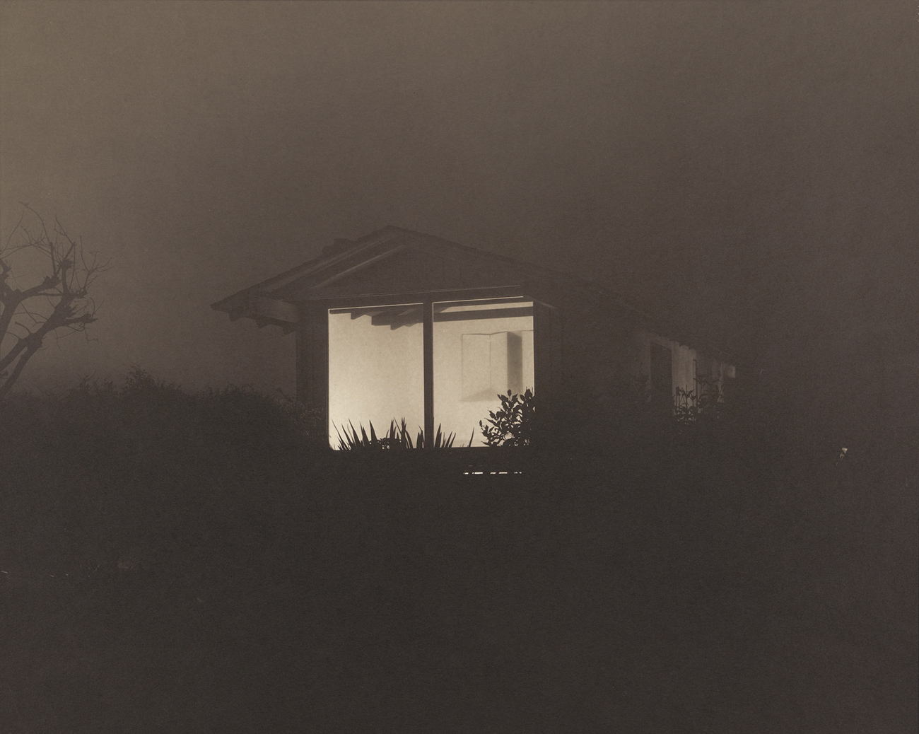 Night time view of a one story structure with a large window looking into an empty lit room.