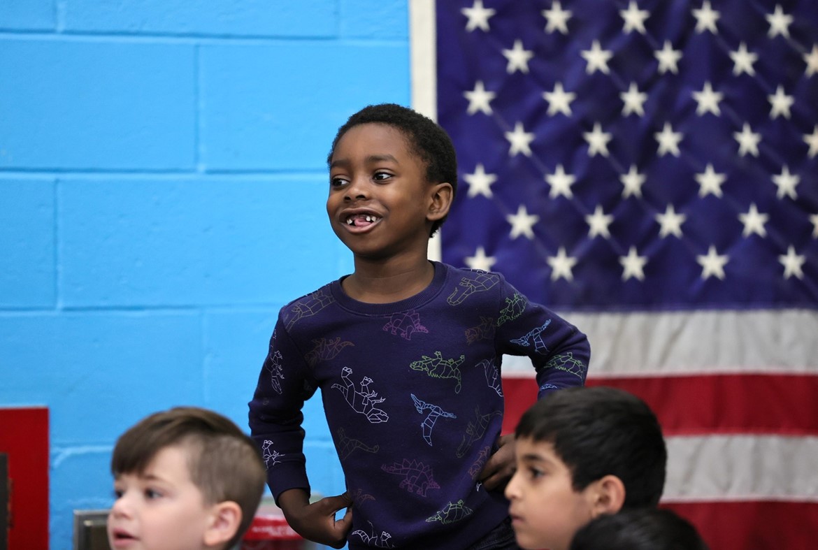 Wood Park kindergartener smiles during spring concert with American flag on wall behind him.