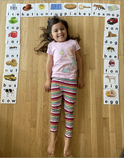A happy Compass Charter School scholar lies in the middle of her spelling homework with three letter words on cards.