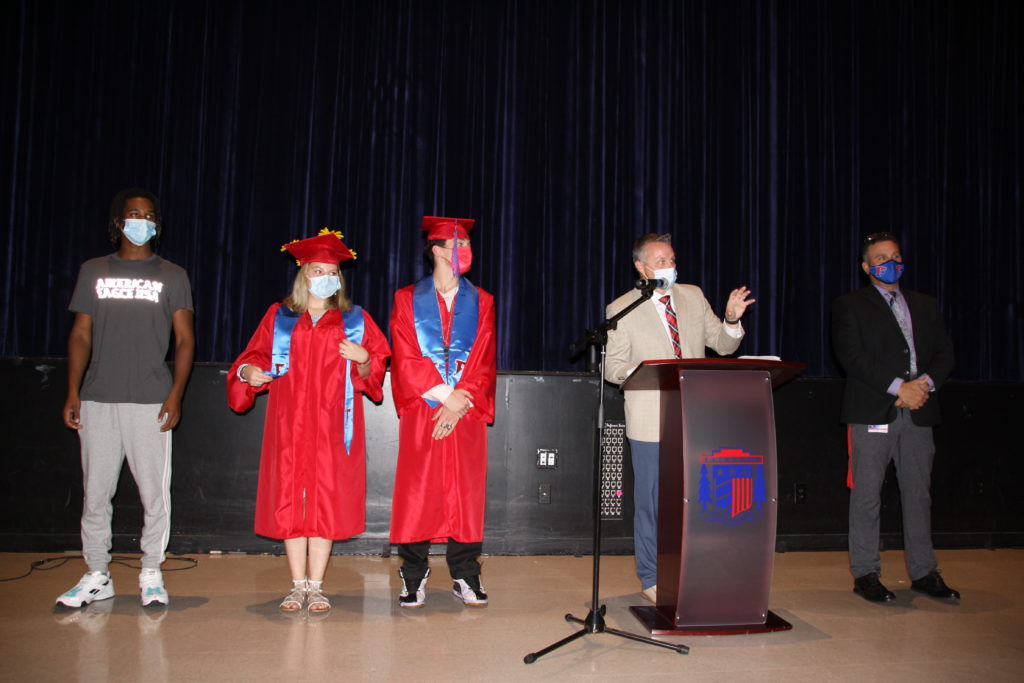 Three students stand on a stage next to a man behind a podium and a second man next to him