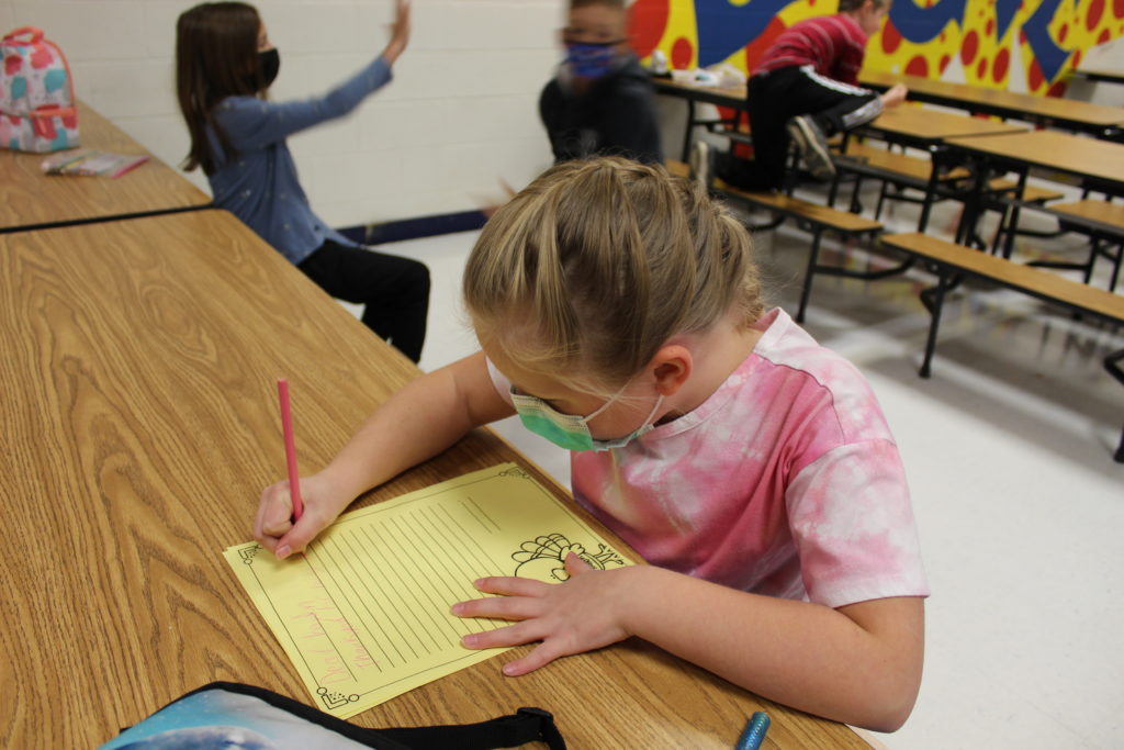 Student in a pink shirt works on a piece of paper at a school lunch table