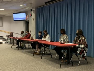 A panel of people sit behind long tables in front of a blue curtain