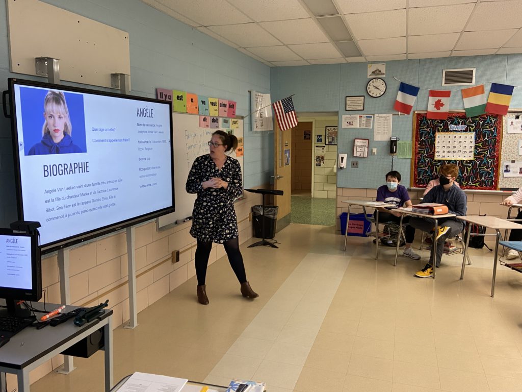 Teacher stands in front of classroom, gesturing at a presentation