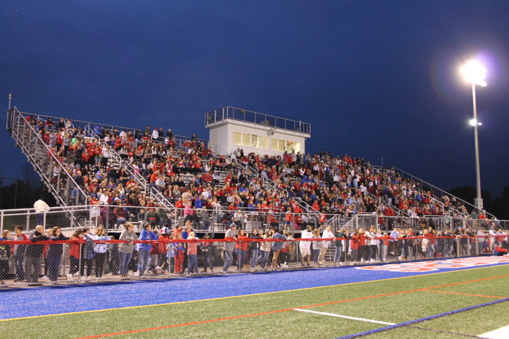 A huge crowd dressed in mostly red fills a football field bleachers