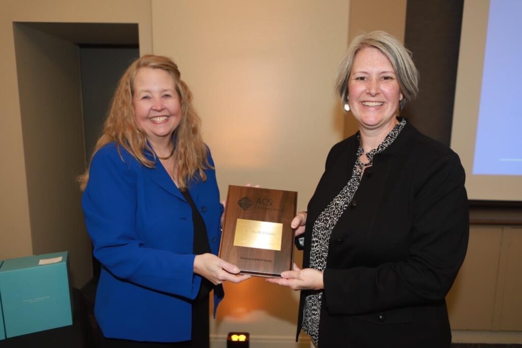 Two women stand on either side of a crystal plaque. Both smile at the camera. The woman on the right is tall with gray hair, the woman on the left is shorter and has red hair.