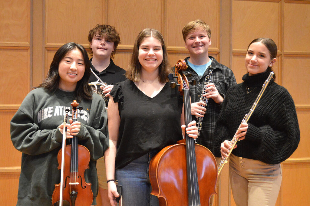 Five students with musical instruments stand in front of a wood panel wall and smile at the camera