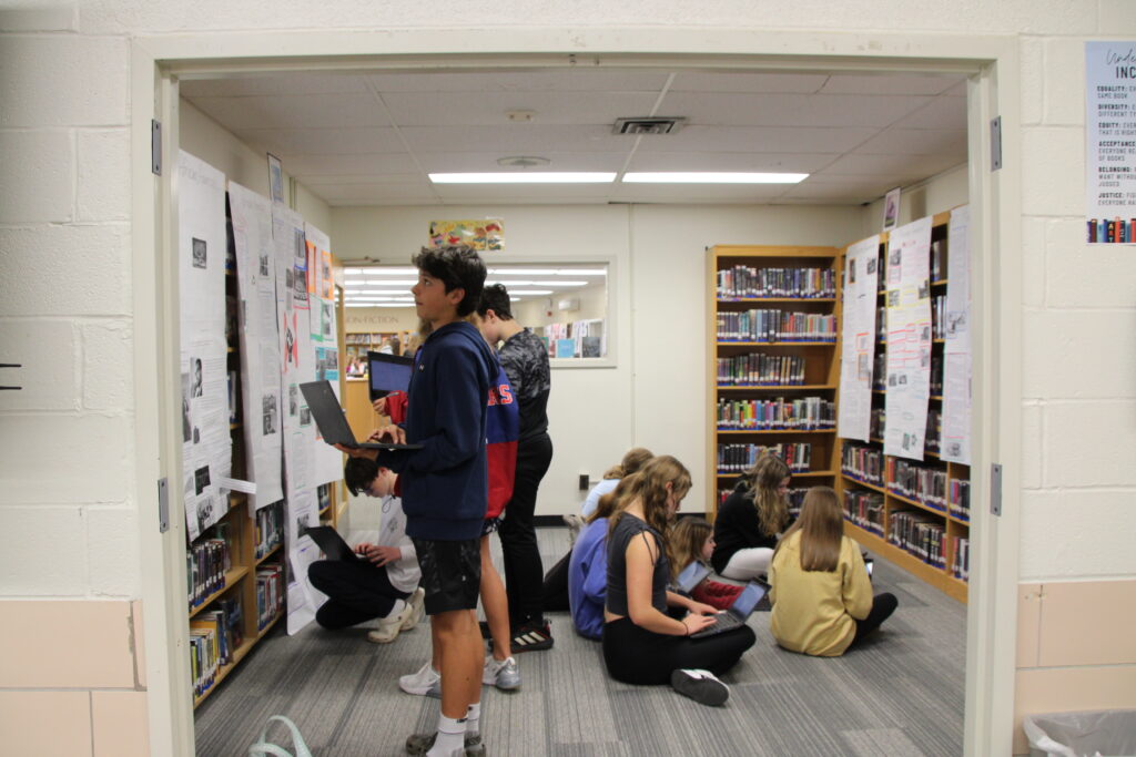 A group of students sit on the floor and look over books while a second group stands and looks at posters taped to library shelves