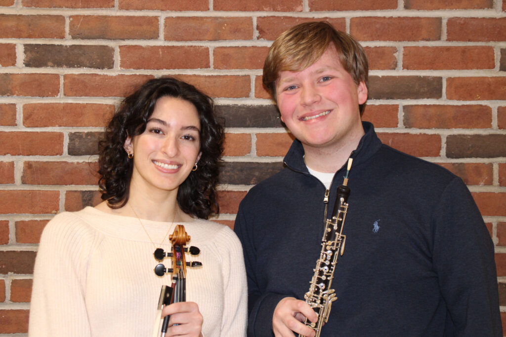 Two high school students stand in front of a brick wall. The student on the left is a young woman with curly dark hair. She is holding the neck of a violin. The student on the right is a young man. He has blond hair and is holding an oboe.