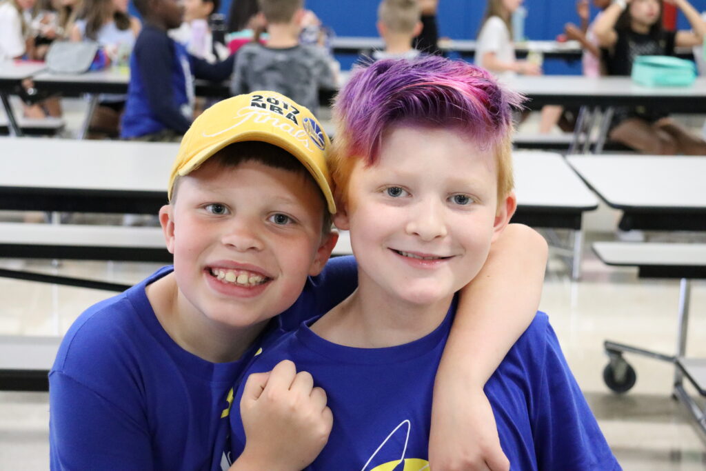 Two elementary school students smile at the camera. One is wearing a yellow hat and the other has bright purple hair