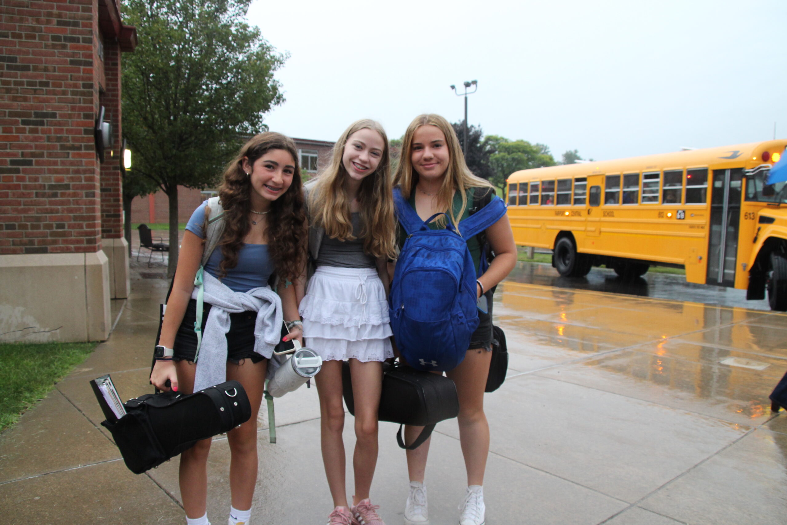 Three middle school students smile at the camera as a school bus drives away behind them