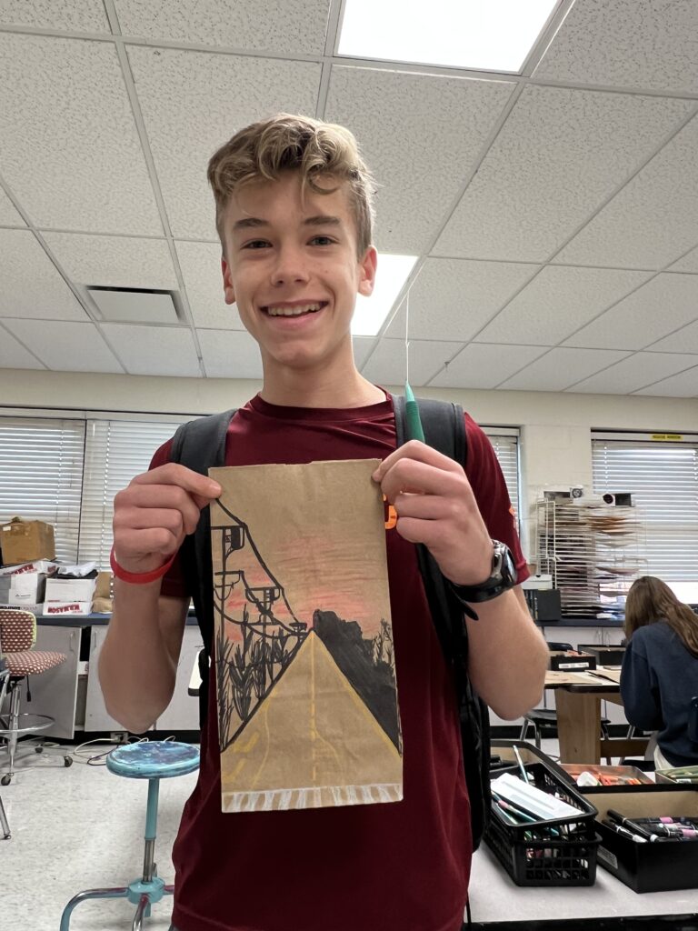 High School student holds up a paper bag on which he has created a scene.