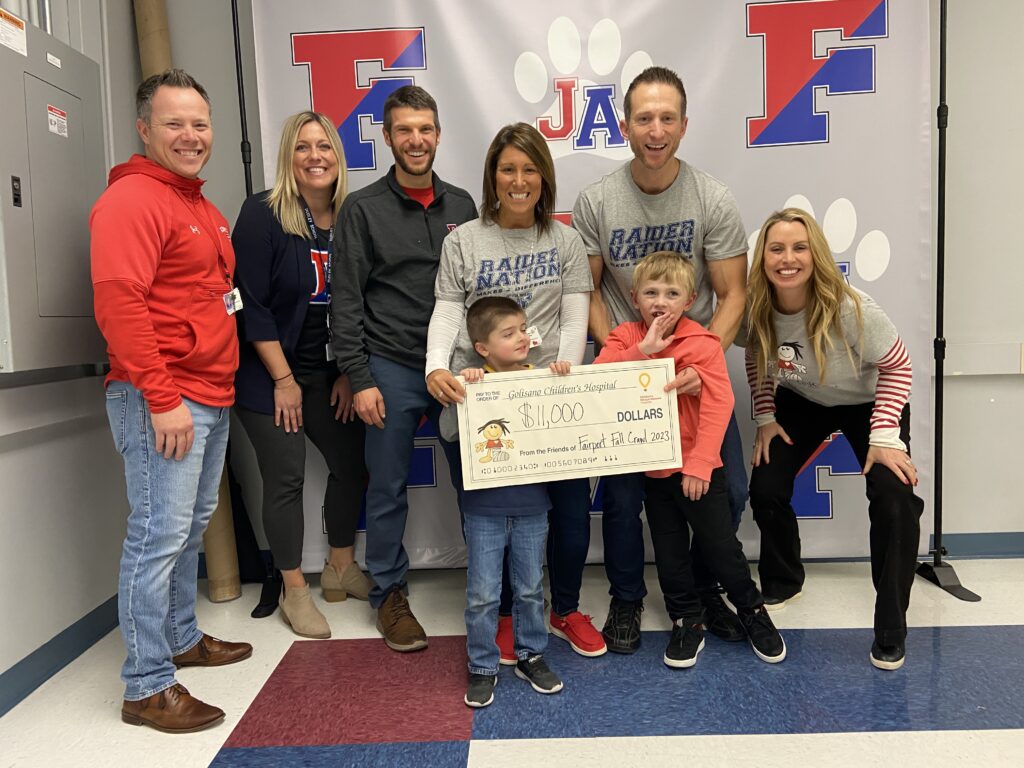 A group of adults stand in front of a photo backdrop. Two young children stand in front of them, holding a large novelty check.