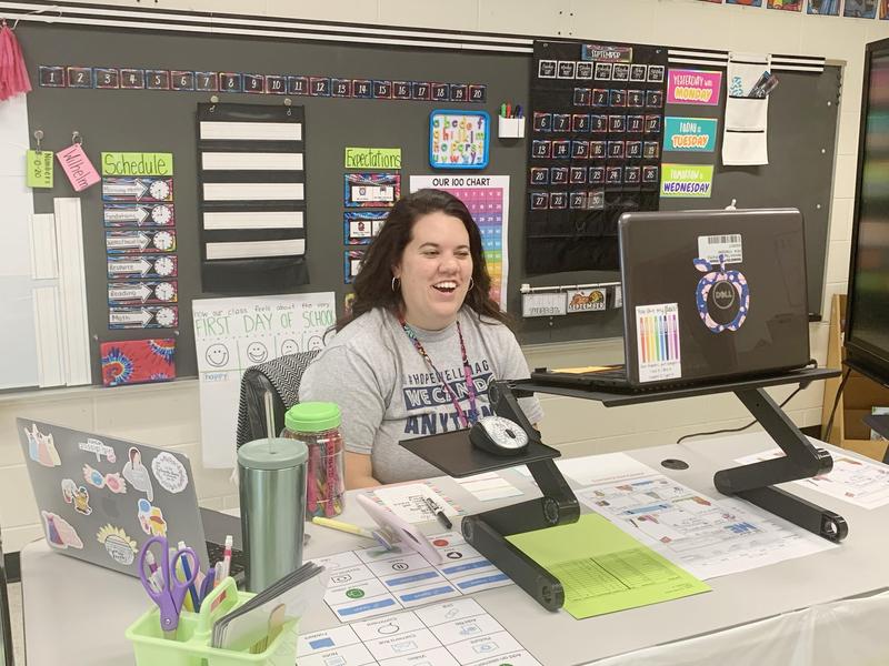 the image is of a teacher wearing a grey shirt sitting at her desk. her laptop is in front of her. she is teaching virtually and smiling.