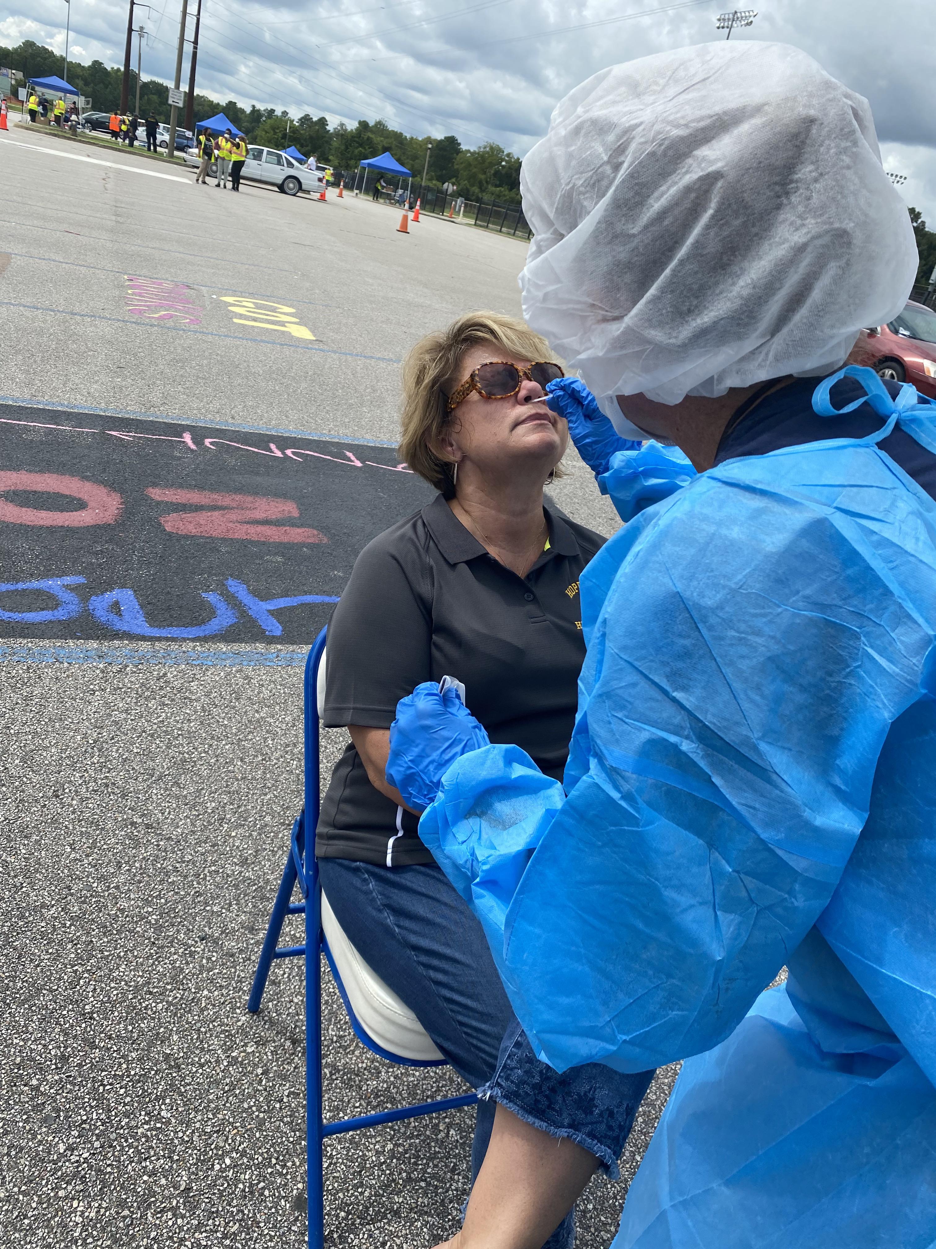 This image is of Dr. Melody Hackney in a black polo shirt and blue jeans. She is sitting on a blue folding chair while a COVID test administrator, in full personal protective equipment (PPE), is swabbing Dr. Hackney’s nose for the test. Image courtesy of HCPS. 