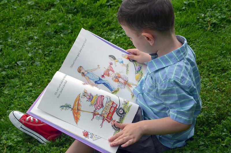 the image is of a young boy in a blue shirt and red sneakers sitting in green grass and reading a picture book