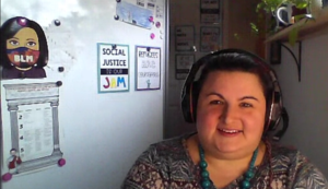 The image is of Ms. Nelson: A kind person with an inviting smile and brown hair. She is wearing large, over-the-ear headphones with an attached microphone and a teal necklace with large spherical beads. On the white wall behind her there are several hangings including a poster that says “Social Justice is my Jam,” a poster that says “Refugees are Courageous,” and a picture of Ms. Nelson’s “bitmoji” wearing Black Lives Matter (BLM) mask. Image courtesy of R.M. Carkhuff