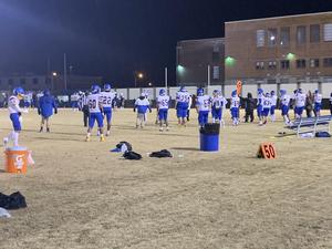 The image is of the varsity football team in their white and blue uniforms. They stand on the sideline of the field. There is an orange gatorade cooler on the ground.