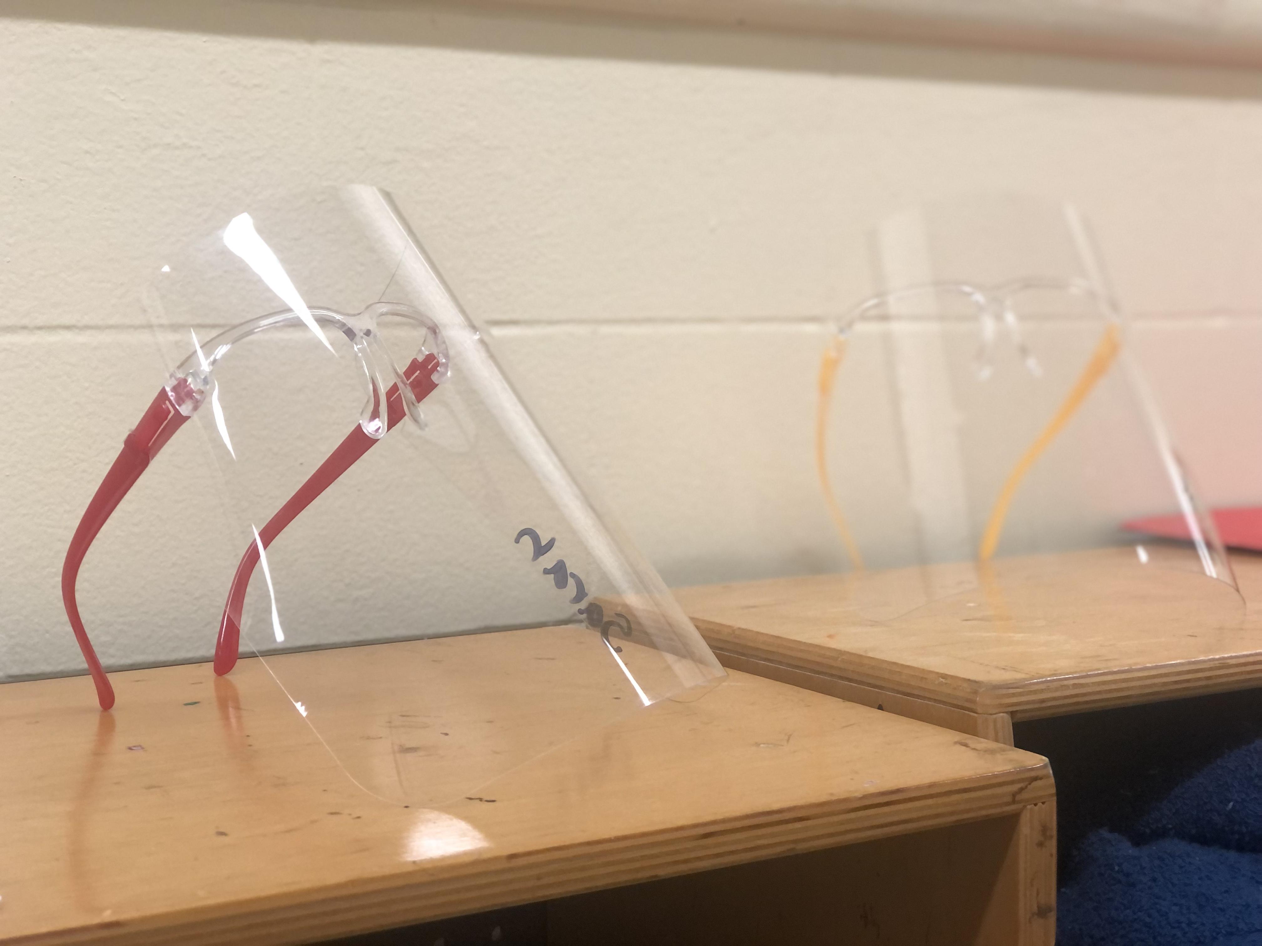 The image is of two glasses-style face shields on top of a wooden cubby. One face shield has red glasses arms and the other has yellow. One face shield has the student’s name down the side. Image courtesy of R. M. Carkhuff. 