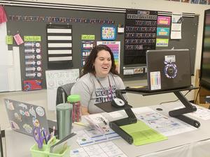 the image is of a teacher wearing a grey shirt sitting at her desk. her laptop is in front of her. she is teaching virtually and smiling.