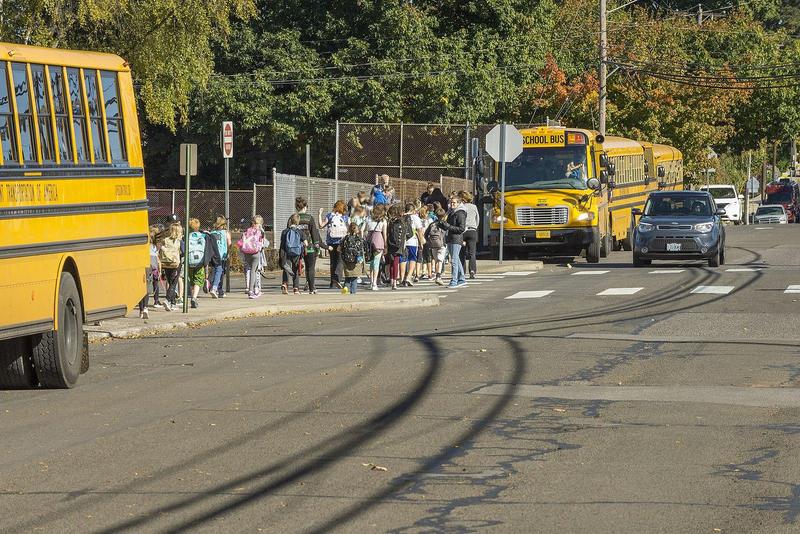 the photo is of a street where 3 school buses are lined up along the sidewalk. A group of students crosses the street with several adults. There is a black car stopped to let the students pass.