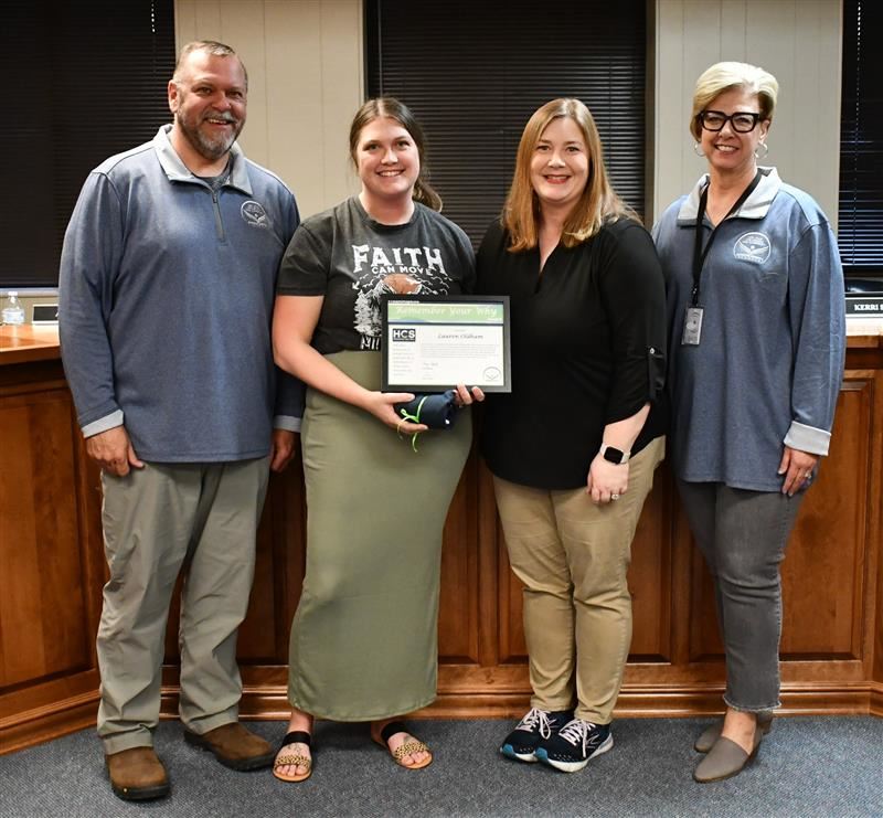 4 people, one holding a certificate, stand in front of desk