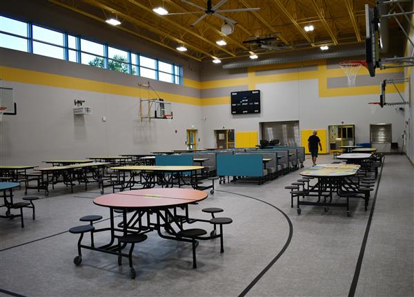 Cafeteria with at least 14 tables and a booth area in the middle, three basketball goals on wall and a scoreboard