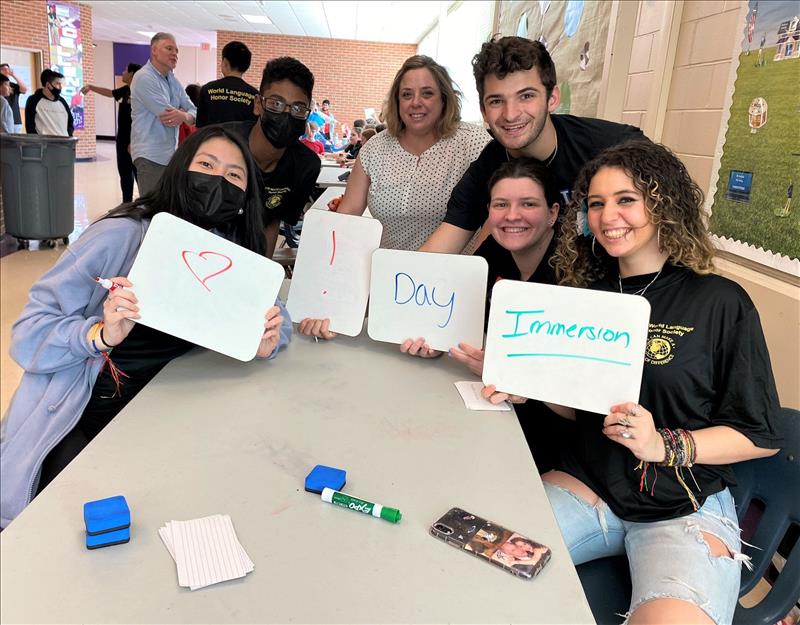 world language honor society students during world language immersion day. Four students are holding white boards one with a heart, one with an exclamation point, one with the word "day" and one with the word "immersion"