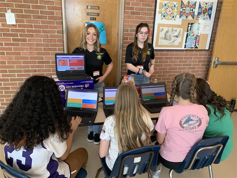 photo of two world language honor society students standing behind a table while four MS students sit at computers facing them