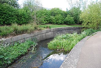 In contrast, a channelized part of the River Ravensbourne, with deliberate gravel shoals and plantings in its bed to make the flow meander and slow it down. This is cheaper than demolishing the concrete (see images below). The adjacent park also acts as a floodplain.[42][43]