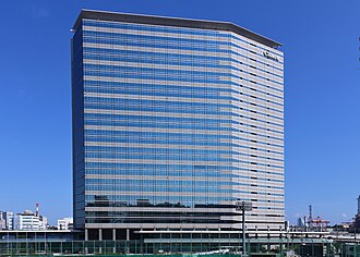 A modern multistorey building in blue and grey color, with Japan Airlines' "JAL" logo on the top right, blue sky on the background, and a highway bridge in the foreground