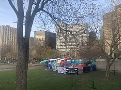 A dense group of tents on a university lawn in downtown Montreal, surrounded by fences bearing banners