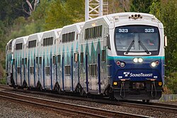 A train with five double-decker passenger cars seen on the furthest of two tracks.