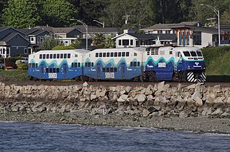 A Sounder trainset, consisting of two double-decker passenger cars and a locomotive, is seen on a track that is slightly elevated over a rocky beach.