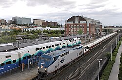 An Amtrak passenger train passes a Sounder train parked on a separate track at a train station with a skyline of mid-rise buildings in the background.