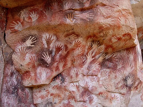 Many painted hand imprints on a rock face in a cave, overlapping