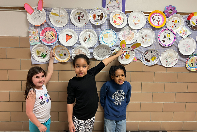 3 young students standing under the wall of paper plates that are decorated