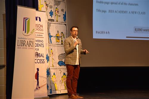 Author standing on stage by a blown up poster of a comic illustration 