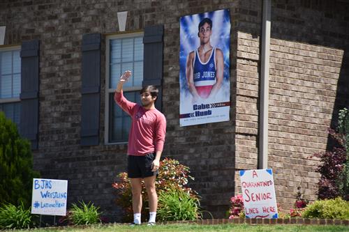 Senior in front yard waving with blowup photo of him as a wrestler for Bob Jones High in background 