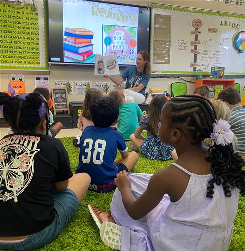 students circled up on floor with teacher reading them a book