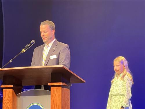 BOE President Tim Holtcamp at podium with student Caroline Turner at his side waiting to lead the Pledge