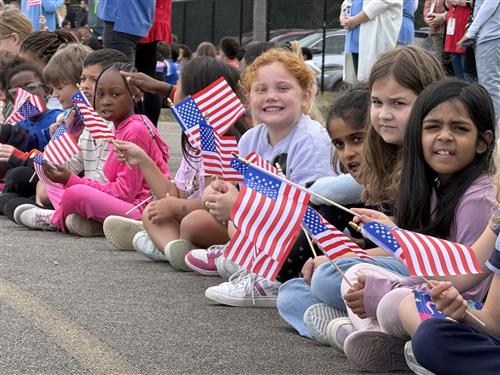 Students lining the curb of a parade route at the school and holding flags
