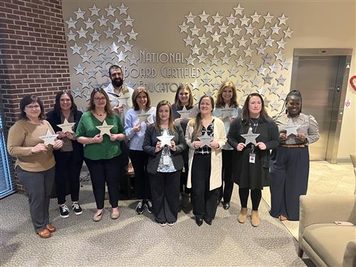 11 new National Board Certified Teachers in Madison City Schools standing with a wall of stars behind them bearing NBCT names